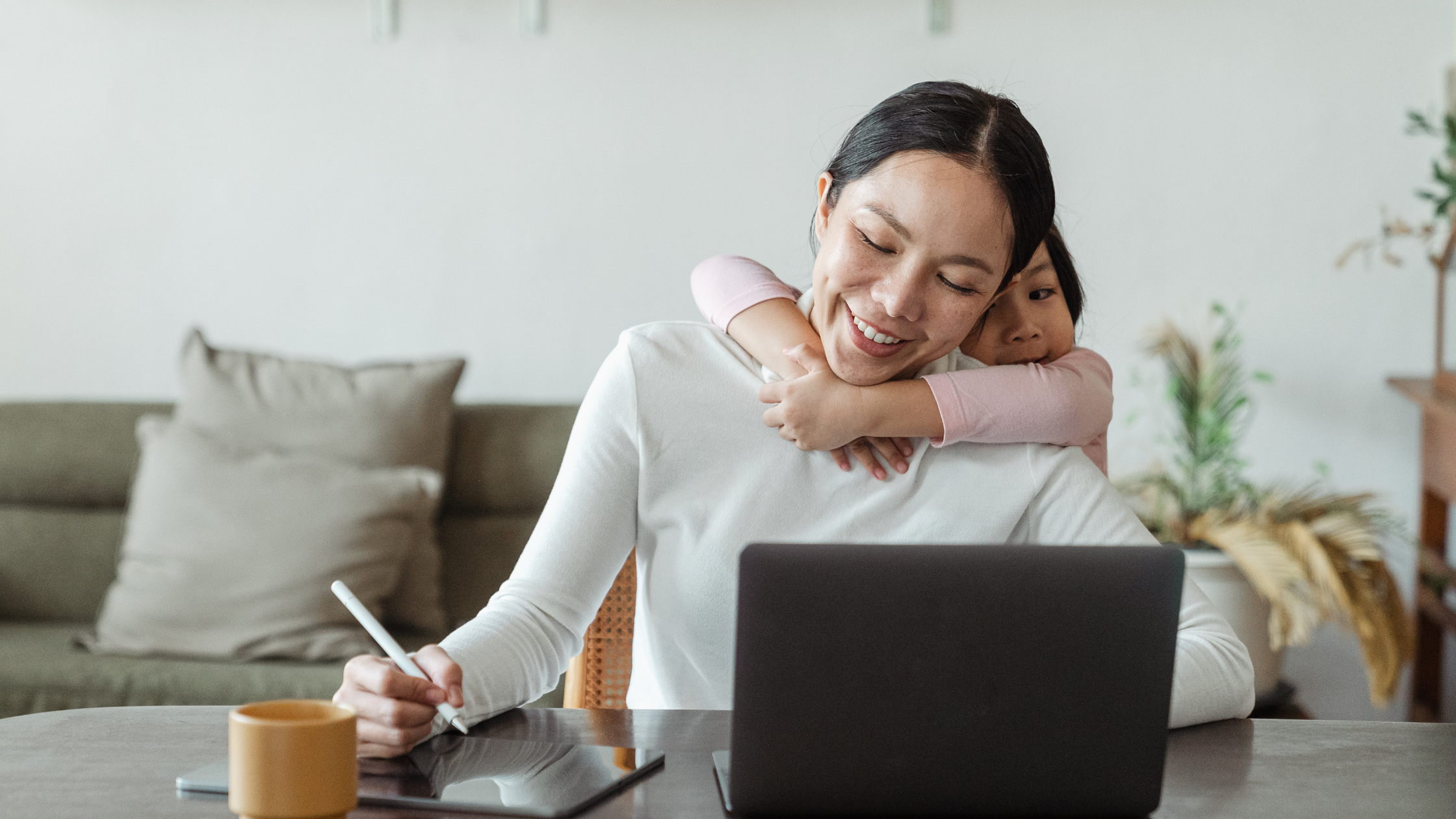 A smiling woman working at a desk with a laptop and writing tablet, while her child lovingly hugs her from behind, depicting the balance of family and pursuing ethical online money-making opportunities