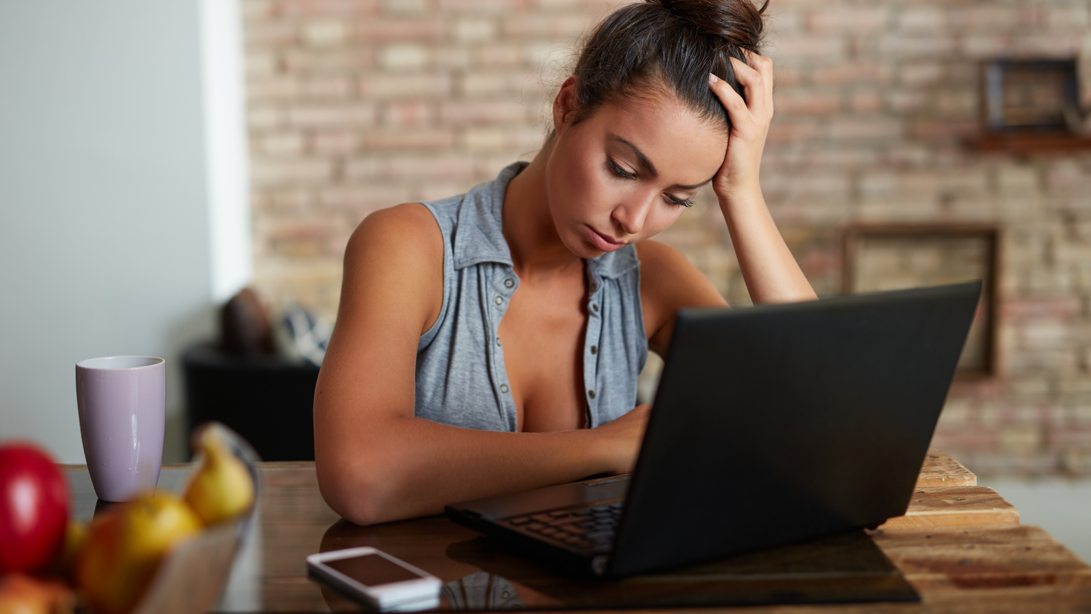 A woman sitting at a table, looking frustrated while working on a laptop. A smartphone and a mug are on the table beside her. This image visually represents the challenges and frustrations faced when trying to make money online ethically, aligning with the blog's theme on ethical and sustainable online income strategies